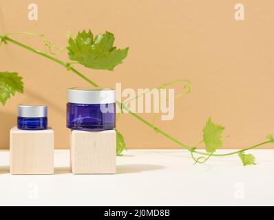 Produits cosmétiques dans un pot en verre bleu avec un couvercle gris sur un podium en bois en cubes, derrière une branche de raisins avec de la lea verte Banque D'Images