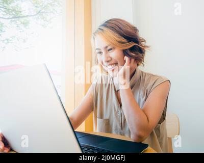 Femme asiatique, freelance avec cheveux courts tendance et regarder l'écran tout en travaillant avec un ordinateur portable sur un bureau rond en bois sur un mur blanc Banque D'Images