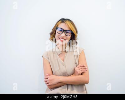 Souriante femme asiatique cheveux courts, style tendance en lunettes noires et chemise sans manches beige posant avec des bras croisés, regardant l'appareil photo sur blanc Banque D'Images