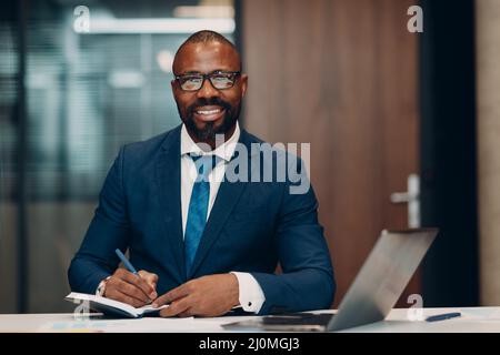 Portrait souriant homme d'affaires afro-américain en costume bleu assis à table pour une réunion au bureau avec un ordinateur portable avec un stylo et un ordinateur portable. Banque D'Images