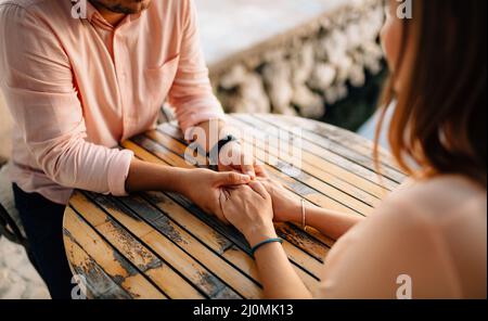 Homme dans une chemise pastel et une montre-bracelet tient les mains de la fille avec des bracelets derrière une table en bois.Gros plan Banque D'Images