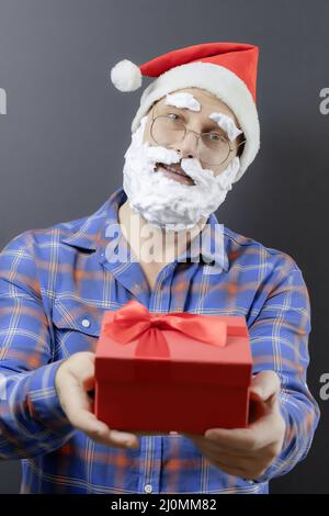 Le père Noël dans une chemise à carreaux avec une barbe en mousse blanche tient une boîte cadeau rouge. Concentrez-vous sur le visage. Joyeux Noël Banque D'Images