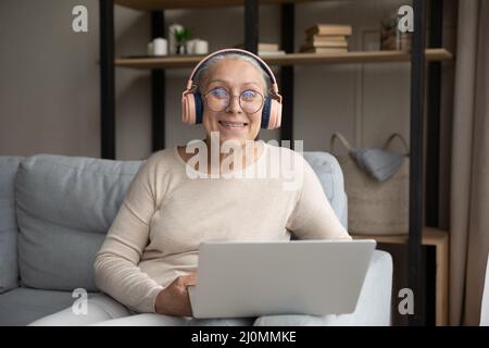 Les écouteurs Happy Senior Wear pour femme sont installés sur un canapé avec un ordinateur portable Banque D'Images