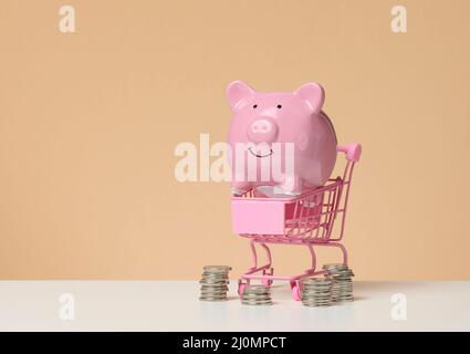 Chariot miniature en métal et banc de porc rose sur une table blanche, fond marron. Concept d'économie de budget, de remises Banque D'Images