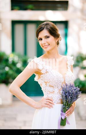 Sibenik, Croatie - 05.06.17: Mariée souriante vêtie d'une robe en dentelle blanche avec un bouquet de stands de lavande reposant sa main sur son côté Banque D'Images