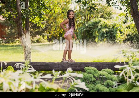 Petite fille jouant en rondins souriant regardant la caméra équilibrant avec des arroseurs d'eau, maison et arbres verts en arrière-plan en journée Banque D'Images