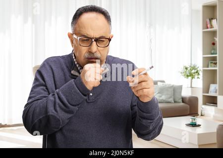 Homme mature fumant une cigarette et toussant à l'intérieur d'un appartement Banque D'Images