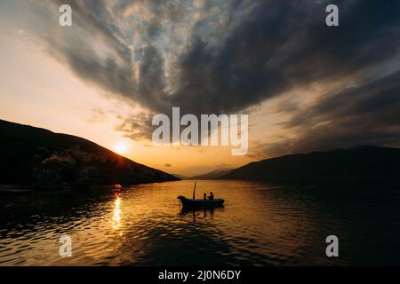 Des silhouettes d'homme et de femme dans un bateau flottent sur l'eau sur fond de montagnes au coucher du soleil Banque D'Images