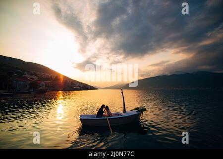 L'homme et la femme s'embrassent dans un bateau au milieu de l'eau sur la toile de fond du coucher du soleil Banque D'Images
