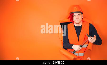 Portrait de jeune homme en casque de protection orange vif et gilet avec un marteau dans ses mains peeking à travers le papier de fond orange déchiré dans studi Banque D'Images
