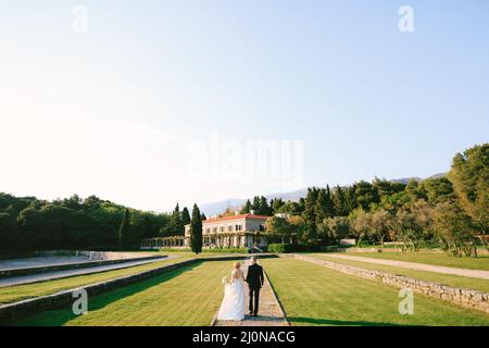 Marié et mariée dans une robe blanche avec un bouquet de fleurs sont en marche le long du chemin dans un beau jardin avec la toile de fond Banque D'Images