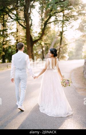 La mariée et marche en tenant les mains sur la route entre les arbres dans une oliveraie, vue arrière Banque D'Images