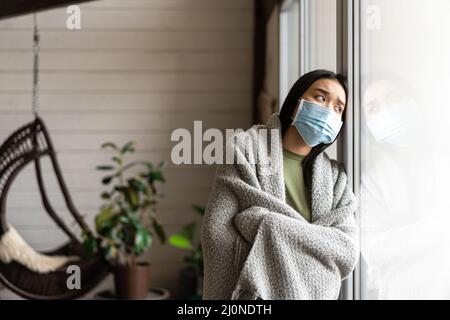 Femme asiatique malade dans un masque médical regardant à l'extérieur de la fenêtre, ayant la grippe ou le covid, rester en quarantaine à la maison Banque D'Images