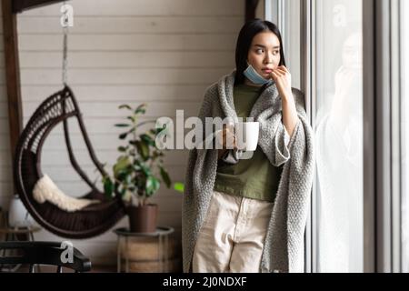 Une jeune femme asiatique boit du thé et regarde à l'extérieur de la fenêtre sur la quarantaine à la maison, étant malade en cas de pandémie de Covid-19 Banque D'Images