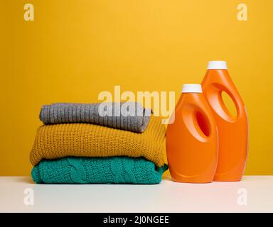 Pile de vêtements pliés lavés et grandes bouteilles en plastique orange avec détergent liquide sur une table blanche, fond jaune Banque D'Images