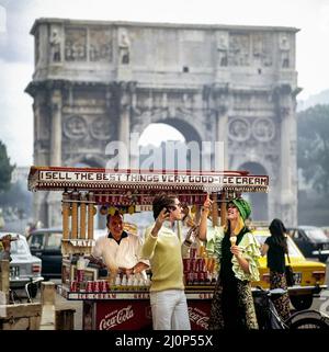 Vintage Rome 1970s, couple s'amusant par rue vendeur de glace en face de l'arc triomphal de Constantine, Italie, Europe, Banque D'Images