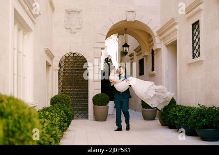 Marié porte mariée dans ses bras dans la cour de la maison avec des arches Banque D'Images