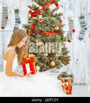 Teenage Girl with long Hair dans une robe Tutu blanche se trouve sous un arbre de Noël et décompresse son cadeau. Le matin de Noël Banque D'Images