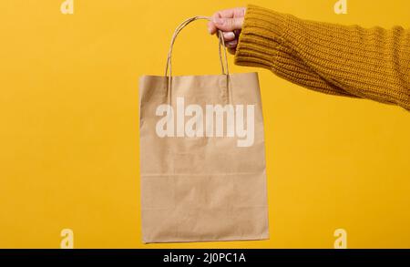 Une main femelle dans un chandail tient un sac de papier marron avec des poignées sur fond jaune. Aucun gaspillage Banque D'Images