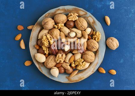 Bleu classique dans la nourriture. Mélange de noix sur l'assiette - noix, amandes, pacanes, macadamia et couteau pour ouvrir la coquille. Nourriture végétalienne saine. Banque D'Images