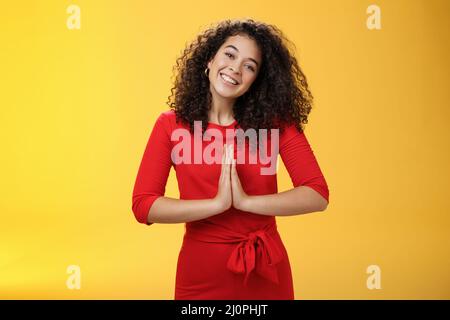 Bienvenue à l'intérieur. Portrait de sympathique et poli bonne-look de la femme hôte en robe rouge avec les cheveux bouclés tenant les mains dans nama Banque D'Images