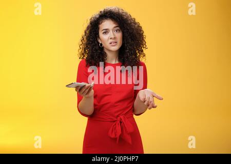 Alors, je me suis embrouillé. Interrogée femme incertaine avec les cheveux bouclés dans la robe rouge haussant en regardant sans indice comme tenir la main ne peut u Banque D'Images