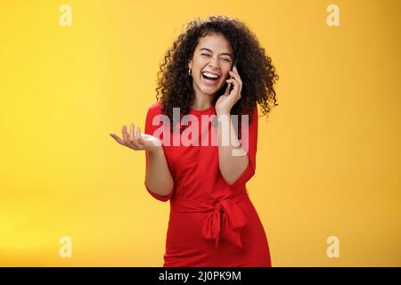 Portrait de joyeuse femme européenne charmante en robe rouge avec des cheveux bouclés riant à voix haute comme des potins avec l'ami via smartpho Banque D'Images