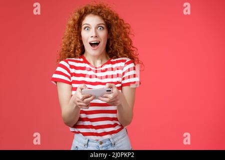 Une étudiante féminine à tête rouge aux cheveux bouclés impressionnée et amusante gagne de manière inattendue un smartphone, un appareil photo, un coup de souffle Banque D'Images