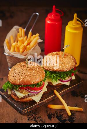 Deux hamburgers faits maison avec du bœuf, du fromage et de la marmelade d'oignons sur une planche de bois, des frites dans un panier métallique et des sauces. Conc. Restauration rapide Banque D'Images