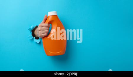 Bouteille en plastique orange avec détergent liquide dans une main femelle sur fond rose. Une partie du corps dépasse d'un trou déchiré Banque D'Images