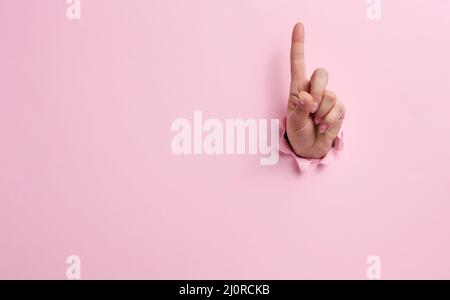 Main féminine qui dépasse d'un trou déchiré sur fond de papier rose, geste d'attention Banque D'Images