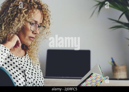 Portrait latéral d'une adorable femme adulte lisant un carnet portant des lunettes. Concept étudiant mûr à la maison avec ordinateur portable en arrière-plan. Chambre simple moderne Banque D'Images