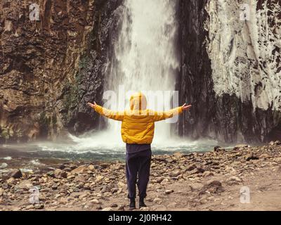 Une femme en blouson jaune se tient avec ses mains à part, regardant une cascade de glace de tempête coulant sur des rochers humides. Banque D'Images