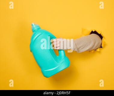 Bouteille en plastique bleu avec détergent liquide dans une main femelle sur fond jaune. Une partie du corps dépasse d'un trou déchiré Banque D'Images