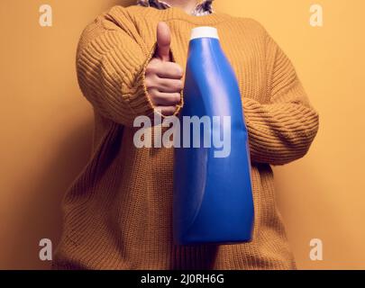 Femme dans un pull tient des bouteilles en plastique bleu avec du gel de lavage liquide. Linge et corvées, fond jaune Banque D'Images