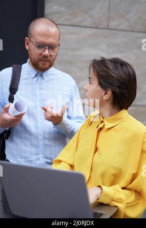 Les collègues de l'équipe d'affaires discutent de se tenir à l'extérieur de l'immeuble de bureaux avec des plans et un ordinateur portable. Collègues concepteurs, agents immobiliers dans les vêtements décontractés brainstorming à l'extérieur. Image de haute qualité Banque D'Images