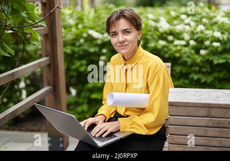 Portrait d'une jeune femme d'affaires heureuse et qualifiée, 30s ans, assise dans le parc avec un ordinateur portable, regardant l'appareil photo. Femme entrepreneur indépendant travaillant à l'extérieur du bâtiment de bureau. Image de haute qualité Banque D'Images