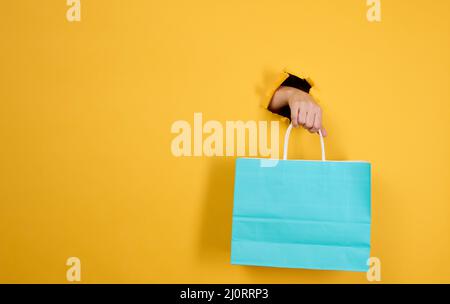 Main féminine avec un sac de shopping en papier bleu qui dépasse d'un trou déchiré sur un fond en papier jaune. Refus du plastique, commencer Banque D'Images