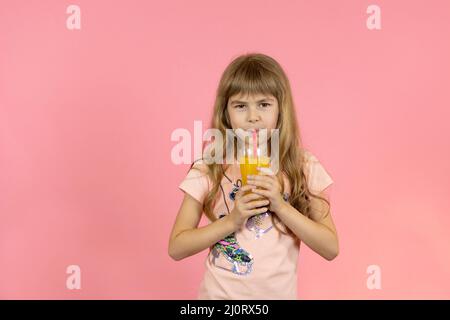 une petite fille boit du jus d'orange sur fond rose. elle tient une orange sur fond rose. Banque D'Images