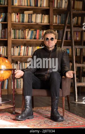 Homme dans une veste en cuir hipster et lunettes de soleil stylées se trouve dans un fauteuil rétro avec des mains ouvertes dans Loft Studio . Étagères à livres bac Banque D'Images