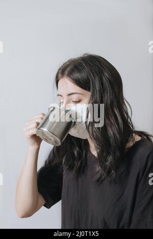 Jeune femme avec une tasse de thé à la fenêtre pendant la quarantaine portant un masque facial inquiet au sujet du nouveau mode de vie. Banque D'Images
