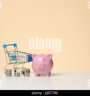 Chariot miniature en métal et banc de porc rose sur une table blanche, fond marron. Concept d'économie de budget, de remises Banque D'Images