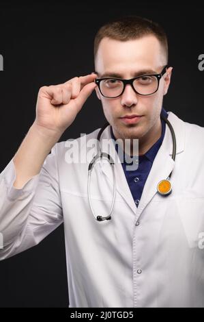 Portrait d'un jeune médecin caucasien en lunettes et en manteau blanc. Permet de tenir les lunettes et de regarder l'appareil photo de manière éreinse Banque D'Images