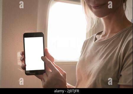 Image de maquette d'une femme tenant et regardant un smartphone avec écran blanc vierge à côté d'une fenêtre d'avion avec des nuages et sk Banque D'Images