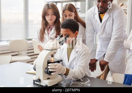 Portrait d'une jeune fille noire regardant au microscope tout en faisant des expériences avec un groupe d'enfants en laboratoire de chimie scolaire Banque D'Images