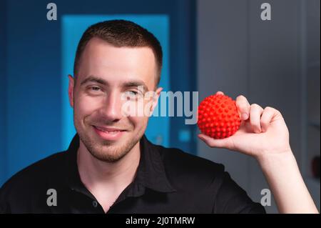 Portrait d'un jeune masseur kinésithérapeute caucasien sur fond bleu avec une boule de massage rouge dans les mains. Souriant et toilettes Banque D'Images