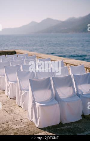 Une rangée de chaises en tissu blanc couvre des supports près d'une clôture en pierre sur le bord de mer Banque D'Images