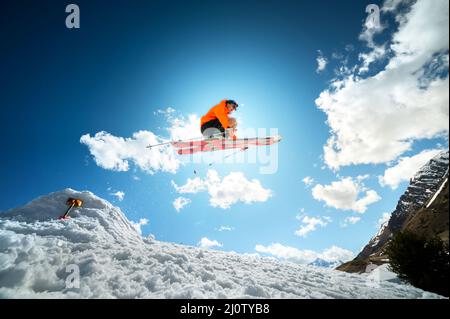 Un jeune homme élégant en lunettes de soleil et une casquette fait un tour en sautant avec un kicker de neige contre le ciel bleu et le soleil o Banque D'Images