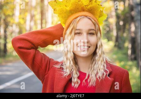 Jolie fille blonde avec des cheveux bouclés dans une casquette orange et un manteau rouge tient un bouquet de feuilles jaunes et sourit contre le roa Banque D'Images