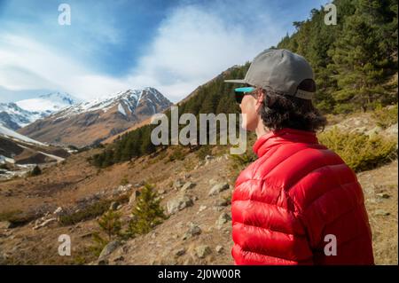 Un jeune homme aux cheveux longs, vêtu d'une veste en duvet rouge et d'une casquette grise en lunettes de soleil, se tient avec son dos à l'appareil photo et regarde une mounta Banque D'Images